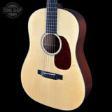 A close-up studio shot of an acoustic guitar against a solid black background, featuring a natural wood top, dark reddish-brown pickguard, and dark fretboard. A Tone Shop watermark is visible in the upper left corner.