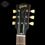 Close-up of a Gibson Les Paul Standard electric guitar headstock, featuring a black face with gold Gibson and Les Paul Standard lettering, cream tuning pegs, and six silver tuning keys, set against a solid black background.