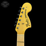 Close-up of a Fender Stratocaster electric guitar headstock in a natural maple finish, showing six silver tuning pegs and the Fender logo, set against a solid black background.