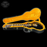 An open black electric guitar hard case with bright orange-yellow velvet lining, holding a black double-cutaway electric guitar. The case is photographed on a solid black background, with a Tone Shop watermark in the upper left corner.