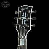 Close-up of a Gibson electric guitar headstock with six silver tuning pegs, a black glossy finish, and a mother-of-pearl style logo inlay, shown against a black background.