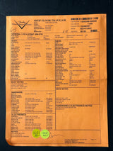 An orange paper shipping and receiving document labeled 'Freddo Custom Mfg' for a 'Shop Floor Traveler', filled with dense printed text and barcodes, photographed on a dark background.