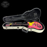 An electric guitar with a pink body and yellow headstock, displayed inside an open white hard case with black lining, set against a solid black background.