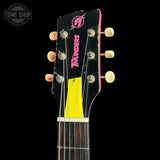 Close-up of a guitar headstock with six cream-colored tuning pegs, a black face, pink 'TRUNDERS' branding, and a bright yellow block near the sound hole, shown against a black background.