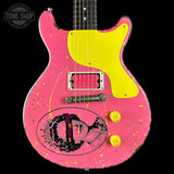 A pink electric guitar with a bright yellow pickguard, yellow knobs, and a distressed finish, featuring a black and white graphic of a woman on the lower left side. The neck is black with white dot inlays.
