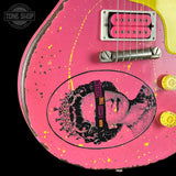 Close-up of a pink electric guitar body with yellow paint splatter, a black-and-pink Queen graphic decal, and part of a yellow headstock visible against a black background. A Tone Shop watermark appears in the top-left corner.