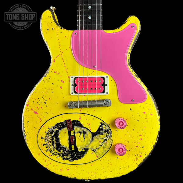 A yellow electric guitar with a distressed finish, featuring a bright pink pickguard, pink pickups, and a black-and-white graphic of a woman's face on the lower bout. The neck is black with white dot inlays.