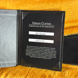 A black Gibson Custom Certificate of Authenticity booklet for an SG Custom instrument, displayed open on a bright yellow textured background.