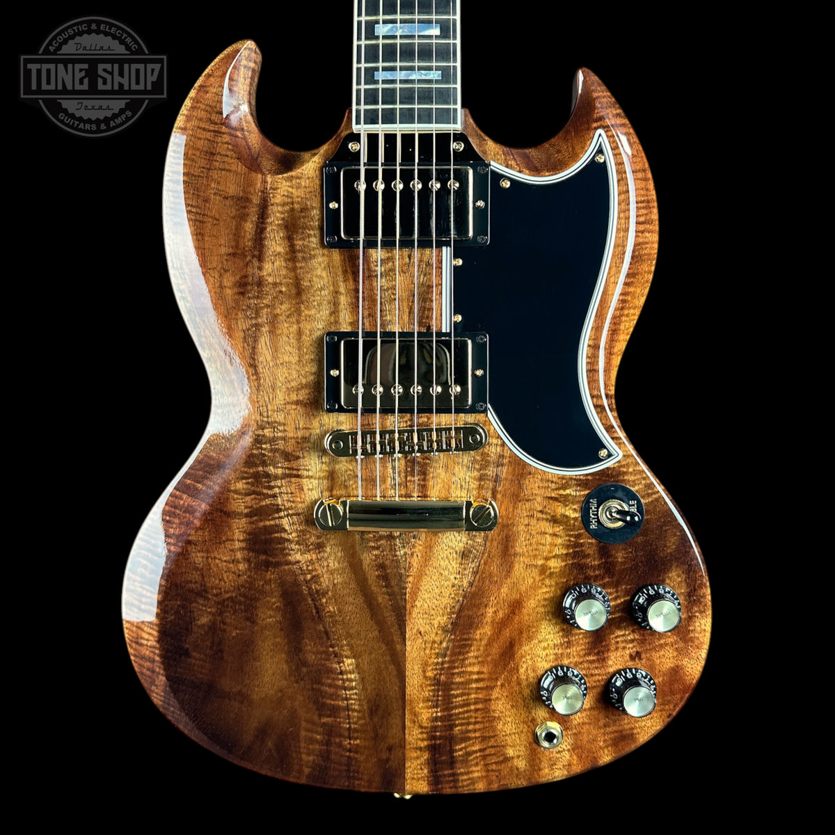A double-cutaway electric guitar with a rich, figured wood finish showing warm brown and golden tones, a black pickguard, and gold hardware, photographed against a solid black background.