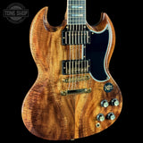 A close-up of a double-cutaway electric guitar with a natural flamed maple wood body, black pickguard, and gold hardware, shown against a solid black background.
