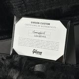 A close-up of a Gibson Custom Certificate of Authenticity card attached to a black ribbon, placed on a black furry surface. The card is white with black text and shows the Gibson logo and Hummingbird model information.