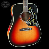 A close-up of an acoustic guitar body showing a sunburst finish that transitions from bright orange-red in the center to dark brown and black around the edges. The headstock is out of frame, but the decorative black pickguard with gold and white floral scrollwork is clearly visible.