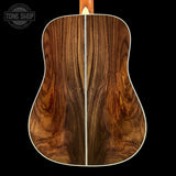 The back of an acoustic guitar made of rich, curly brown wood, featuring a white binding edge and a white ruler placed vertically down the center to show the scale. The headstock is not visible.