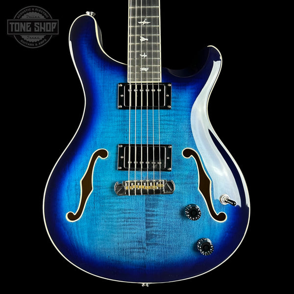 Blue electric guitar with a black pickguard on a black background, featuring 'Tone Shop' branding.