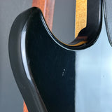 Close-up of a glossy black electric guitar body with a cutaway edge and a light wood neck visible in the background.