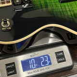 A close-up of a black electric guitar body resting on a digital scale, showing the scale display reading 10.23 oz and part of the guitar's cream binding and green-grained wood top.