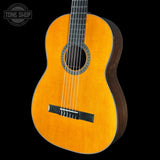 A classical acoustic guitar with a natural wood finish and dark sides, shown against a solid black background with a Tone Shop watermark in the top left corner.