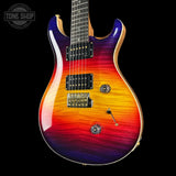 A double-cutaway electric guitar with a vibrant flame-top finish transitioning from purple and blue at the upper shoulder, through orange and yellow, to red on the lower bout. The guitar features a dark fretboard, gold hardware, and is photographed against a solid black background with a Tone Shop watermark in the top-left corner.