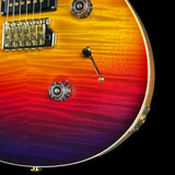 Close-up of a guitar body showing a vibrant flame-tinted finish that transitions from bright yellow and orange to red, purple, and deep blue, with gold hardware.