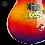 Close-up of a double-cutaway electric guitar body showing a vibrant flame maple finish with a red, orange, yellow, and purple sunburst gradient. The guitar features a black humbucker pickup with gold hardware, a gold bridge, and a chrome control knob, set against a solid black background with a Tone Shop watermark in the top left corner.