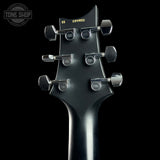 Close-up of the back of a black electric guitar headstock with six silver tuning pegs and yellow serial number printing, shown against a black background with a Tone Shop watermark.