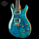 A double-cutaway electric guitar with a vibrant turquoise and teal flamed maple top, featuring gold hardware and a natural wood neck, shown against a solid black background.