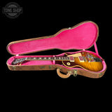 A Gibson Les Paul Custom electric guitar in a sunburst finish, displayed inside its open hard case with a bright pink velvet interior. The guitar is positioned horizontally, and the brand logo is visible on the headstock.