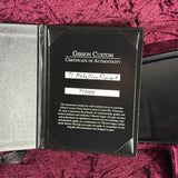 A Gibson Custom Certificate of Authenticity booklet opened to show the black insert with handwritten serial details, placed on a textured magenta velvet surface.