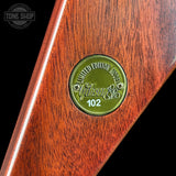 Close-up of a Gibson 102 limited edition headstock inlaid with reddish-brown wood, featuring a circular green metal emblem with the Gibson logo and 'Limited Edition 102' text.