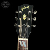 Close-up of a Gibson acoustic guitar headstock against a black background, showing the gold Gibson logo, faceted tuning pegs, strings, and part of the fretboard.