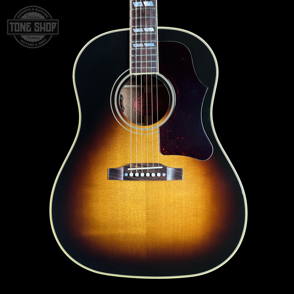 A classic acoustic guitar with a natural sunburst finish, featuring a dark brown pickguard and a white binding, shown against a solid black background with a Tone Shop watermark in the top left corner.