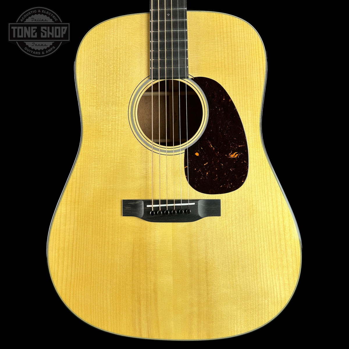 A close-up studio shot of an acoustic guitar's front body, showing a natural wood finish, sound hole, pickguard, and bridge against a solid black background. A Tone Shop watermark is visible in the top-left corner.