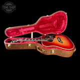 A sunburst acoustic guitar resting inside an open brown hard case with a bright pink velvet interior, shown against a solid black background. A Tone Shop watermark is visible in the top-left corner.