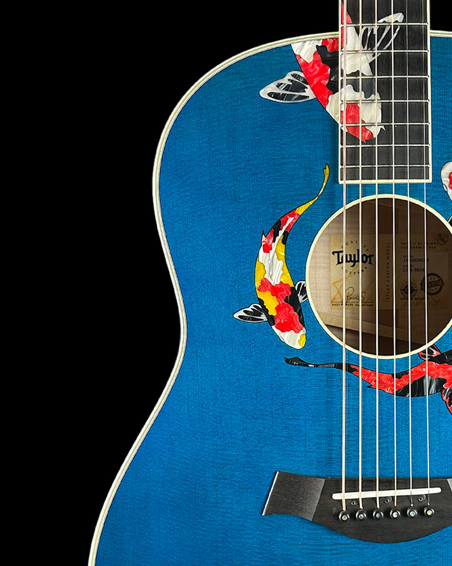 Close up of blue Taylor acoustic with inlayed koi fish around soundhole and fretboard against a black background