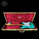 Teal electric guitar in a gold case with red interior, brand logo visible.