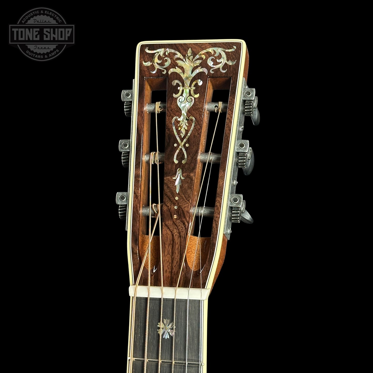 Close-up of a guitar headstock with intricate design on a black background, featuring the Tone Shop logo.